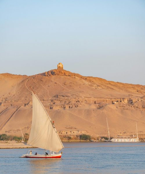 A sailboat on the Nile River with desert hills and ancient structures in the background.