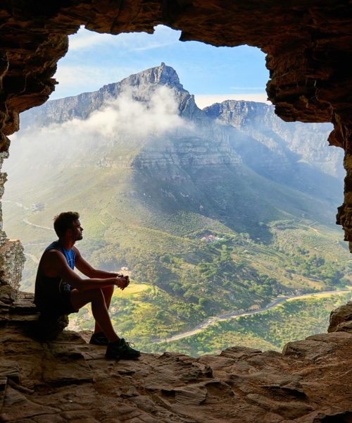 A man sitting in a cave overlooking a majestic mountain landscape under daylight.