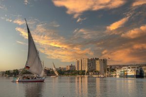 A traditional felucca sails the Nile at sunset with cityscape in the background.